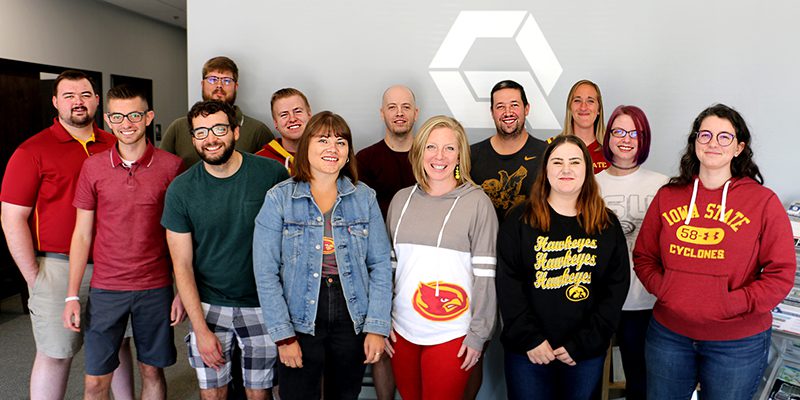 A group of 14 smiling people stands indoors in front of a wall with a geometric logo. Most are casually dressed in T-shirts and sweatshirts, some with Iowa State and Iowa Hawkeyes logos.