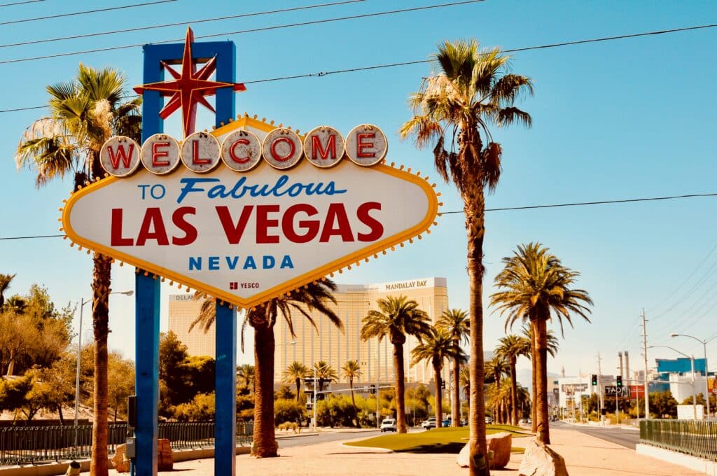 The iconic “Welcome to Fabulous Las Vegas, Nevada” sign stands surrounded by palm trees under a clear blue sky, with city buildings and the Mandalay Bay hotel visible in the background.